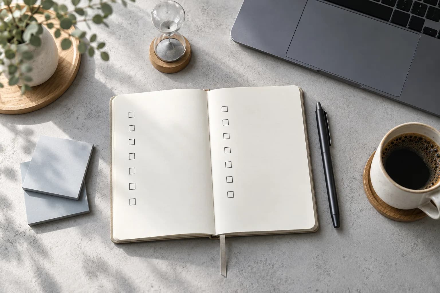 Top-down view of an open notebook with empty checkboxes, a pen, sticky notes, a small hourglass and a coffee cup on a concrete desk