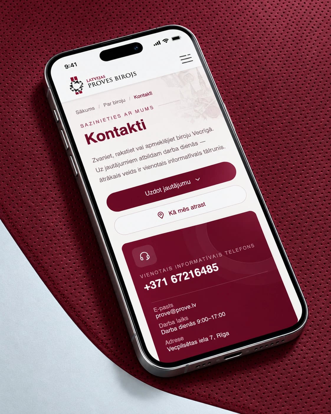 The prove.lv contacts page on a smartphone on a burgundy perforated surface, viewed from above.
